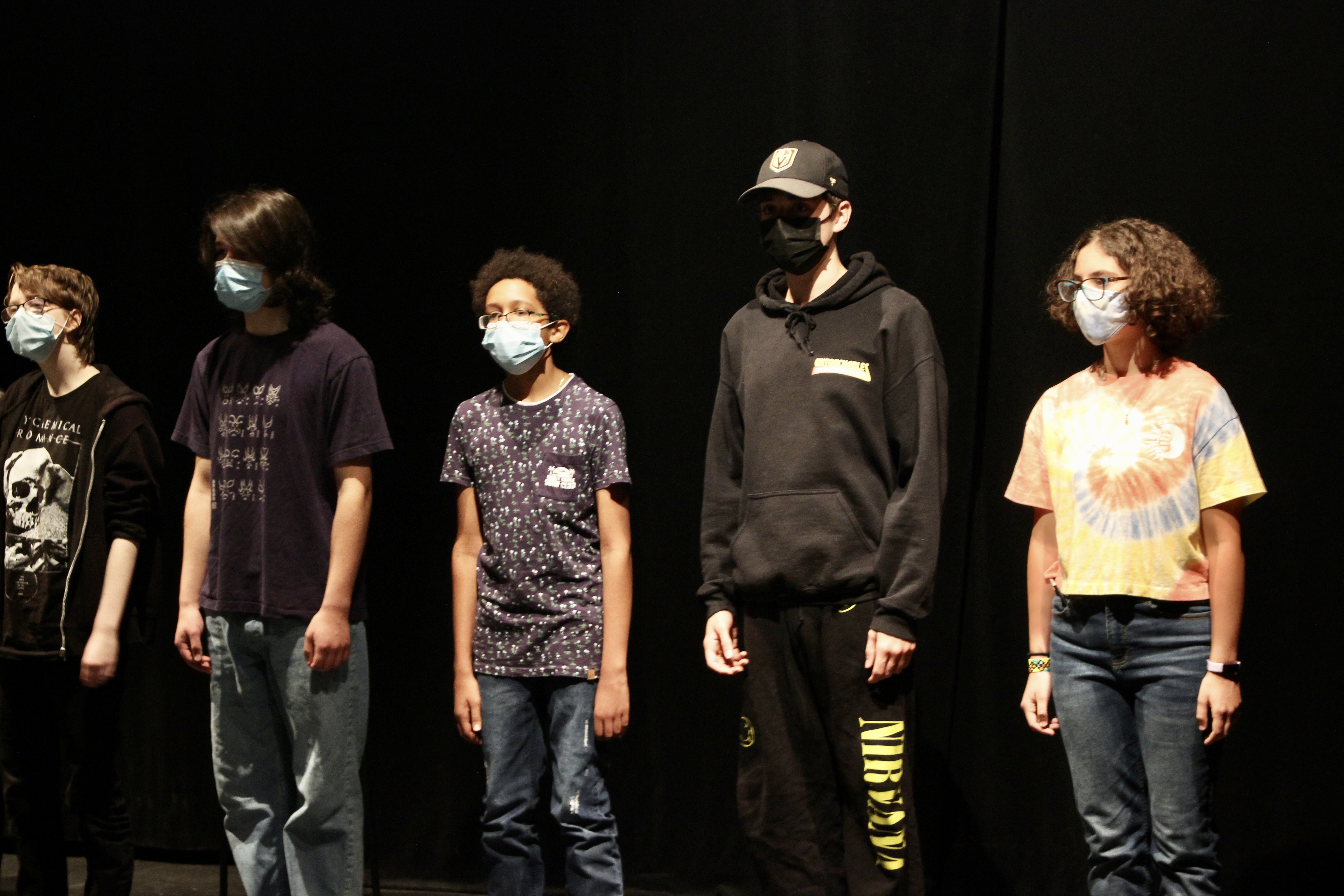 6 youth wearing causal clothing stand in a line, They wear masks. There is a black background.