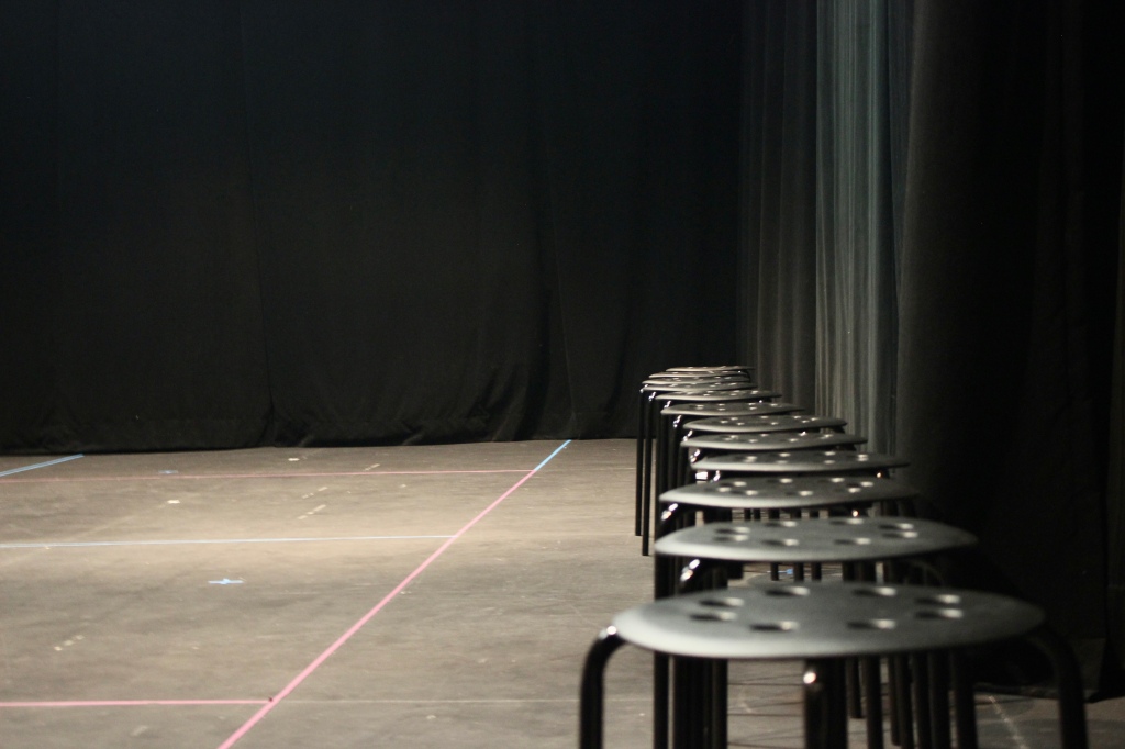 12 black stools in a row inside a theatre studio with a black curtain and dark grey floor