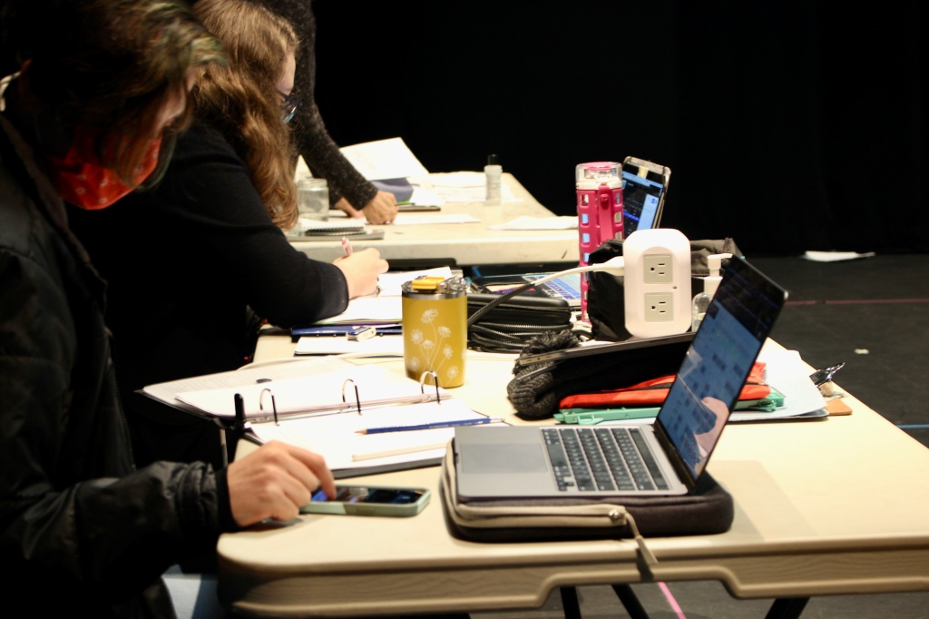 a white long table with books, a lap top, water bottle and other necessities; at the table, two women work and take notes